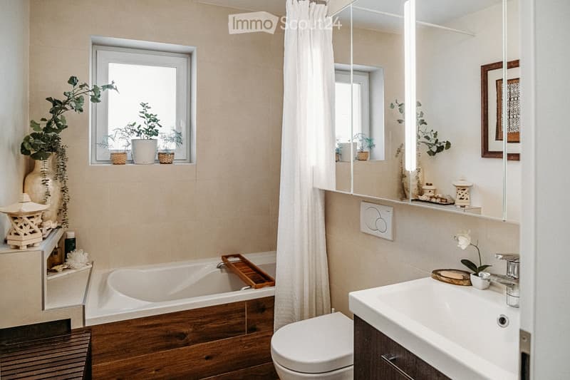 Modern bathroom with a bathtub, toilet, sink, and mirror. Features include a wooden tray on the bathtub, a vase with plants on the windowsill, and a decorative wooden cabinet.