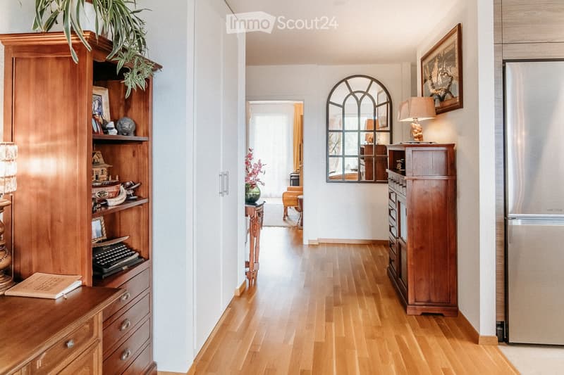 Elegant hallway with wooden flooring, white walls, and a large arched mirror. Features include a wooden cabinet with decorative items, a small table with a vase of flowers, and a refrigerator on the right side.