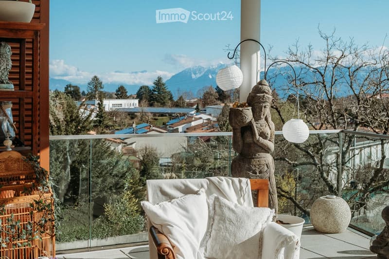 Balcony with a view of mountains, featuring a wooden chair with white cushions, a decorative statue, a hanging lantern, and a ceramic vase.