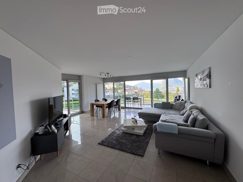 Modern living room with large windows offering a scenic view, featuring a grey sectional sofa, coffee table, side table, dining table, and chairs.