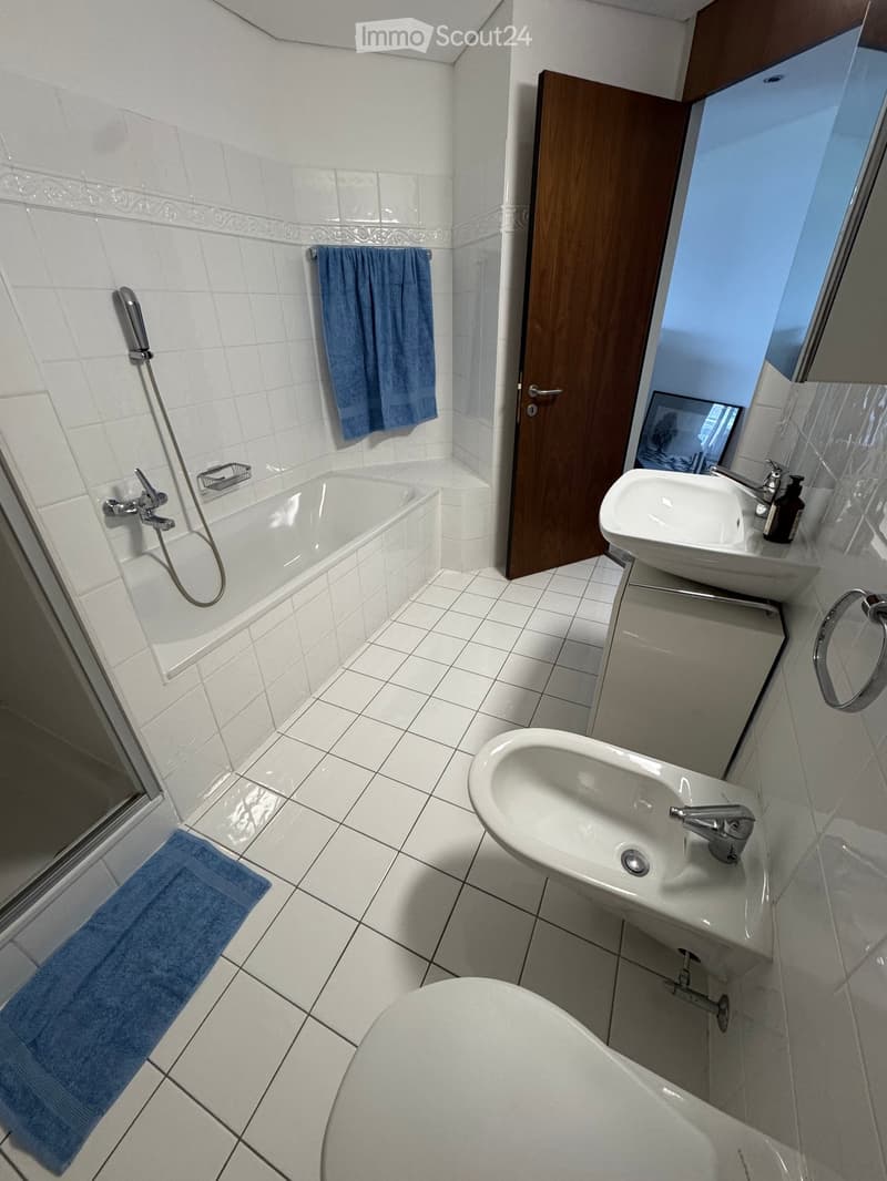 Modern bathroom with white tiled walls and floor, featuring a bathtub, sink, toilet, and towel.