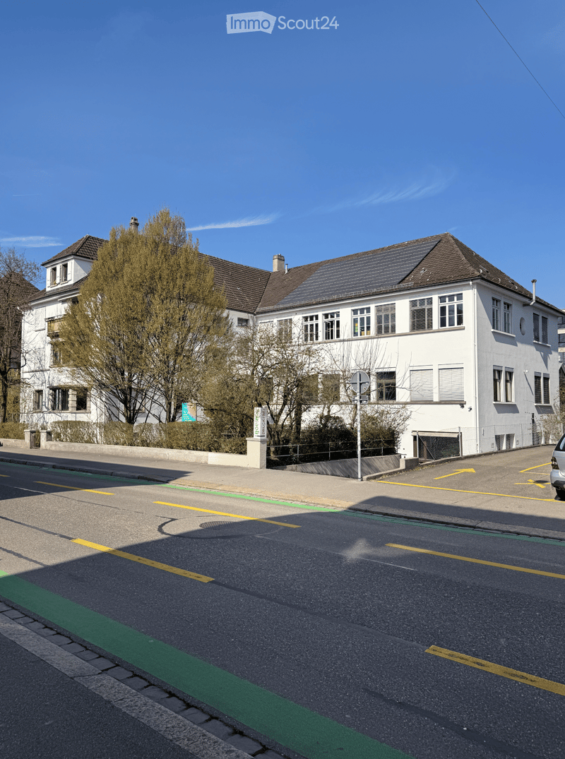 Two-story white building with multiple windows and a brown roof, featuring solar panels on the roof and surrounded by trees and a paved road.