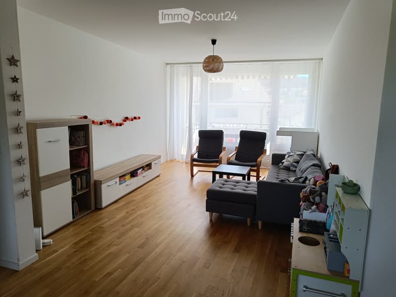Bright living room with wooden flooring, large window with white curtains, grey sofa with ottoman, two armchairs, coffee table, bookcase, and children's play area.