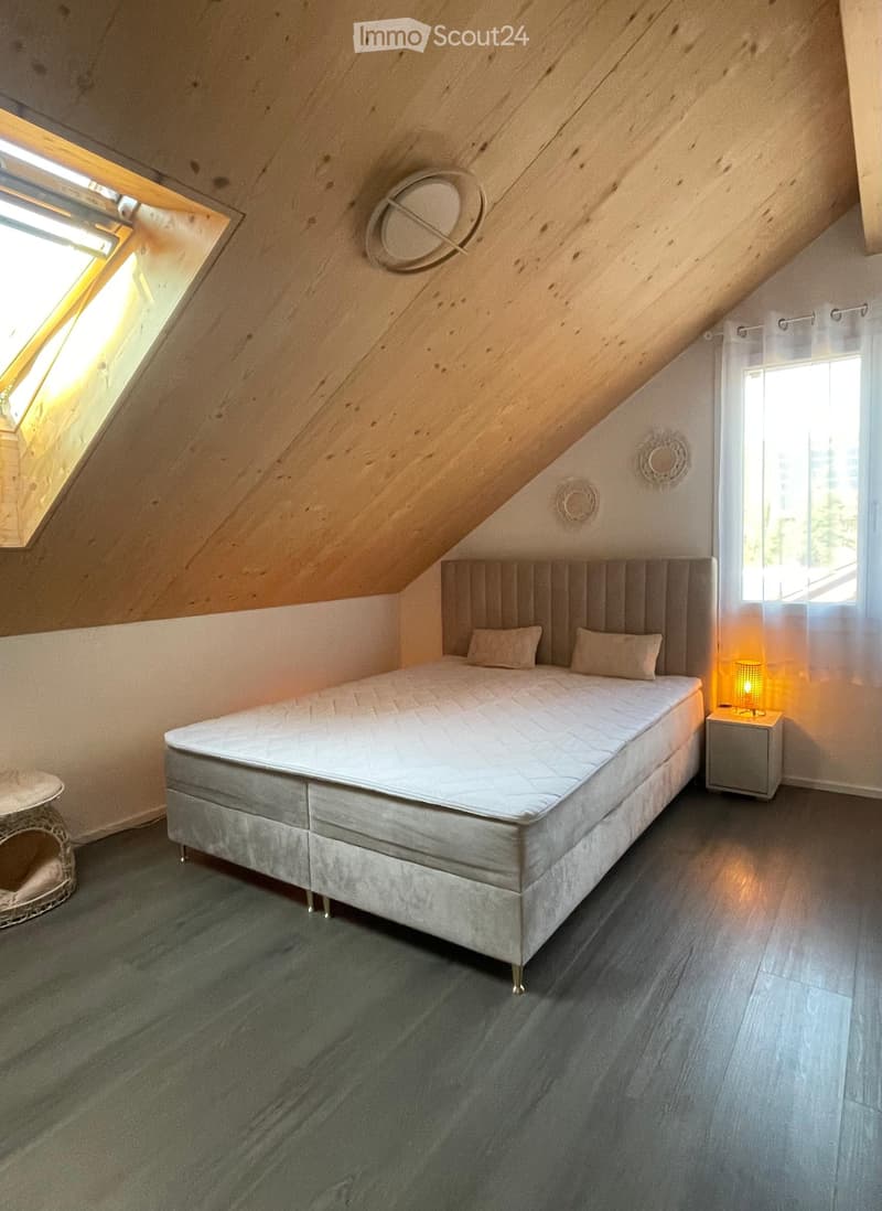Cozy attic bedroom with a large bed, nightstand, table lamp, and wicker basket. The room features wooden slanted ceilings and large windows allowing natural light.