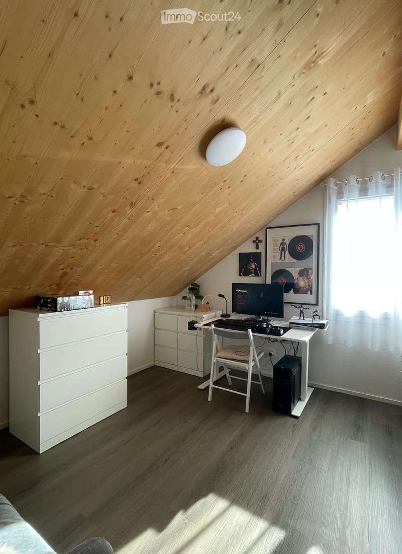 A cozy attic room with wooden slanted ceilings, a desk setup with a computer, chair, and accessories, a white cabinet, and a small plant on the windowsill.