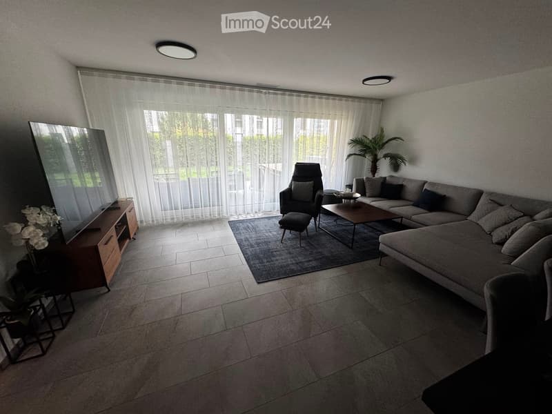 Modern living room with large windows, grey sectional sofa, coffee table, armchair, ottoman, TV stand, and decorative plant.