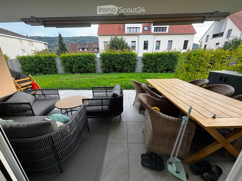 Balcony with a sofa, small table, chairs, and a larger wooden dining table with chairs, surrounded by greenery and a view of neighboring houses.
