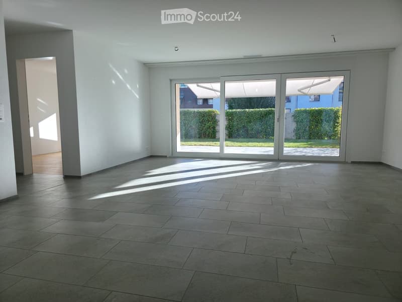 Empty room with tiled floor, white walls, large sliding glass doors leading to a garden, and natural light streaming in.