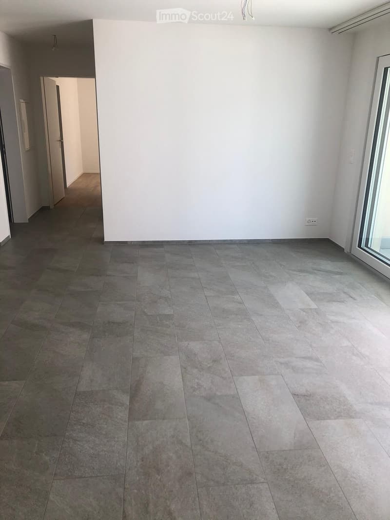 Empty room with grey tiled floor, white walls, and a large window on the right side. An open doorway leads to another room with wooden flooring.