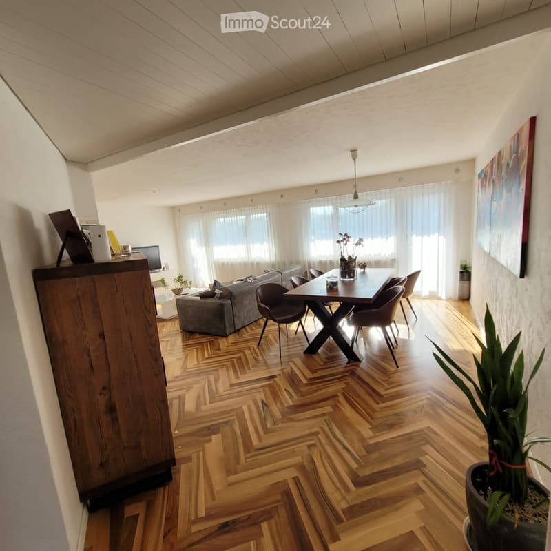 Modern living and dining area with herringbone wooden flooring, large windows with sheer curtains, a grey sofa with ottoman, a wooden dining table with four chairs, and a potted plant near the window.
