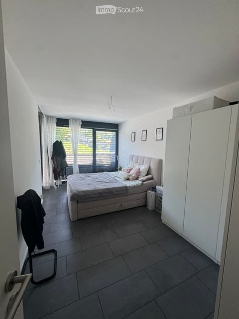Modern bedroom with a large bed, white wardrobe, nightstand, and large window with curtains. The room features dark tiled flooring and several picture frames on the wall.
