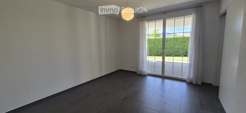 Empty room with wooden flooring, white walls, large sliding glass doors leading to a garden, and a hanging light fixture.