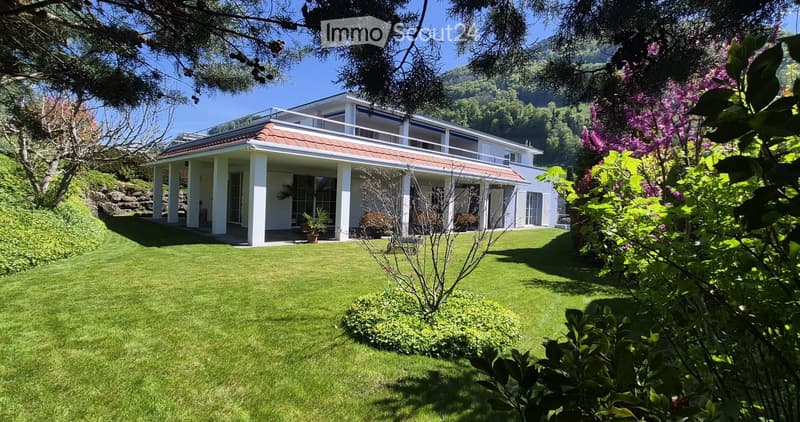 A well-maintained lawn with a modern two-story house featuring a red-tiled roof and white columns, surrounded by lush greenery and flowering trees.