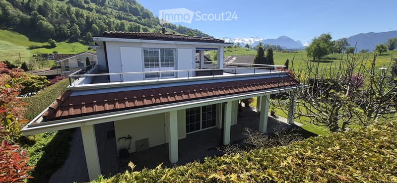 Two-story house with a flat roof terrace, surrounded by greenery and mountains in the background.