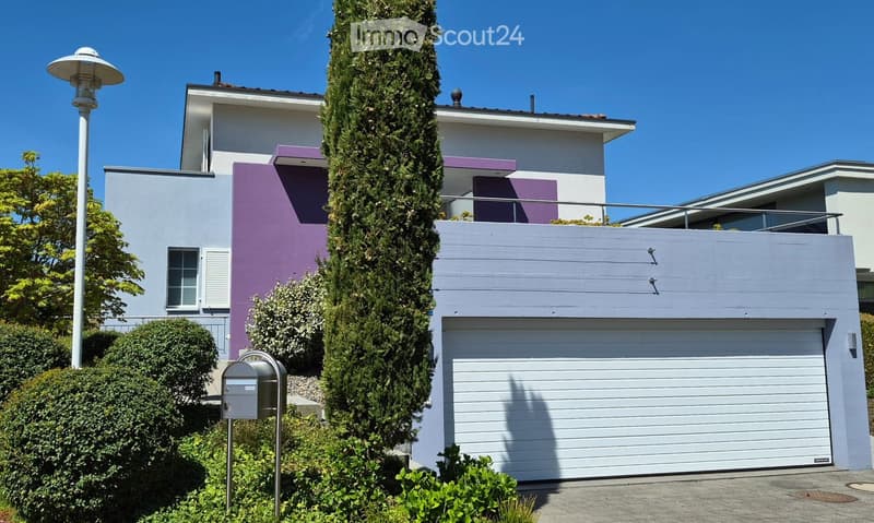 Modern two-story house with white and purple exterior, large garage, and well-maintained landscaping including a tall evergreen tree and shrubs.