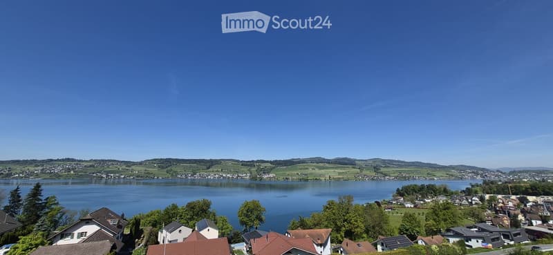 Scenic view of a lake surrounded by green hills and scattered houses with red-tiled roofs, under a clear blue sky.