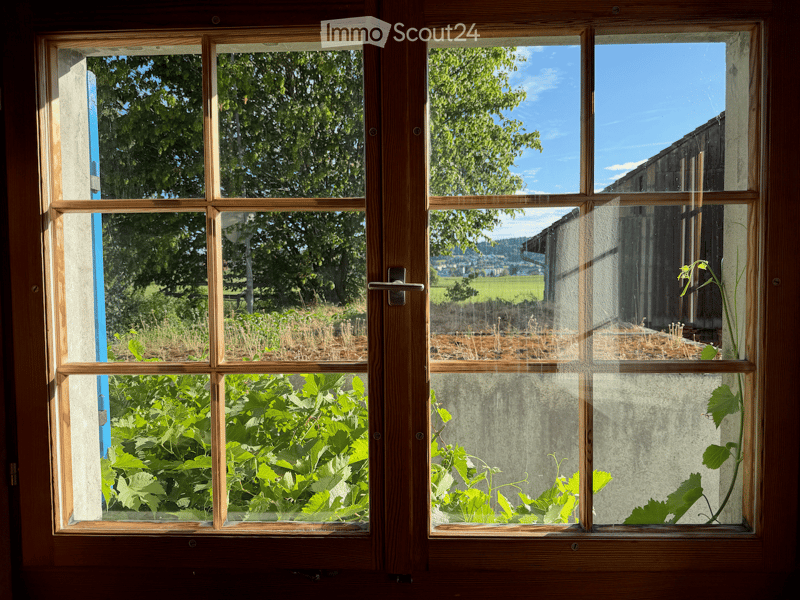 An unfurnished room with large wooden-framed windows showing a view of a garden and countryside landscape. The windows are open, revealing lush greenery and a clear blue sky.