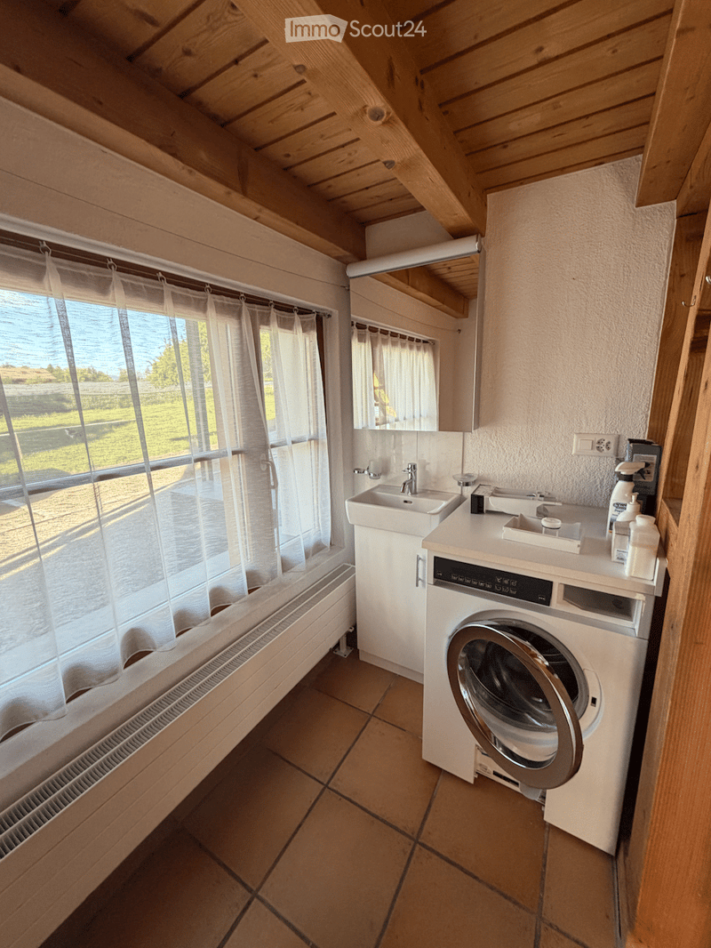 Bathroom with wooden ceiling, tiled floor, sink, washing machine, and large window with curtains.