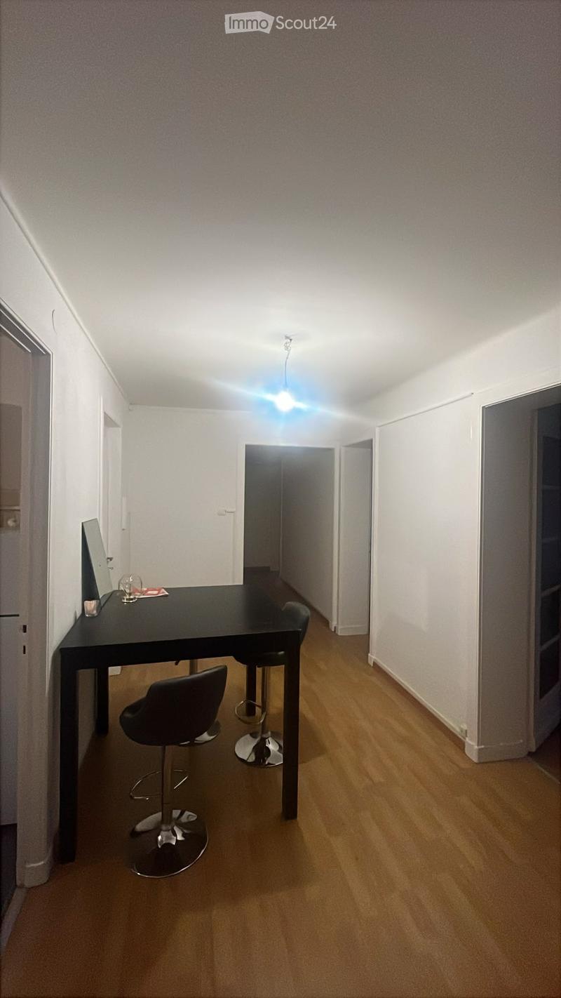 A small, well-lit room with wooden flooring, a black table, two chairs, and two stools. The room has white walls and a ceiling light fixture.