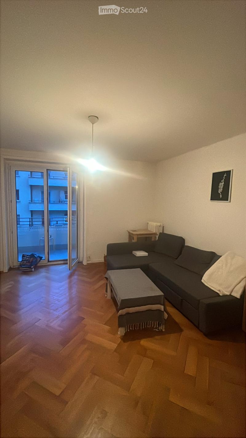 Modern living room with wooden flooring, a dark grey sectional sofa, a coffee table with an ottoman, a small wooden side table, and a radiator near the balcony door.