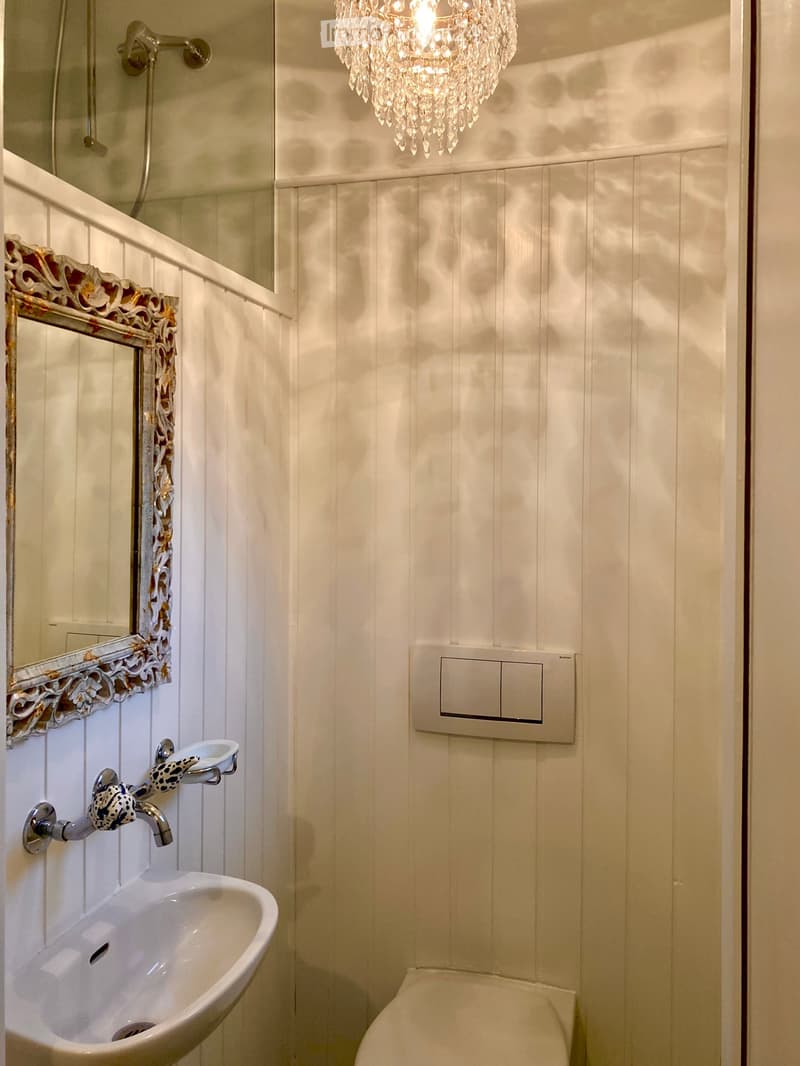 Elegant bathroom with white paneled walls, a crystal chandelier, a sink with a decorative faucet, a toilet, and a mirror with ornate gold detailing.