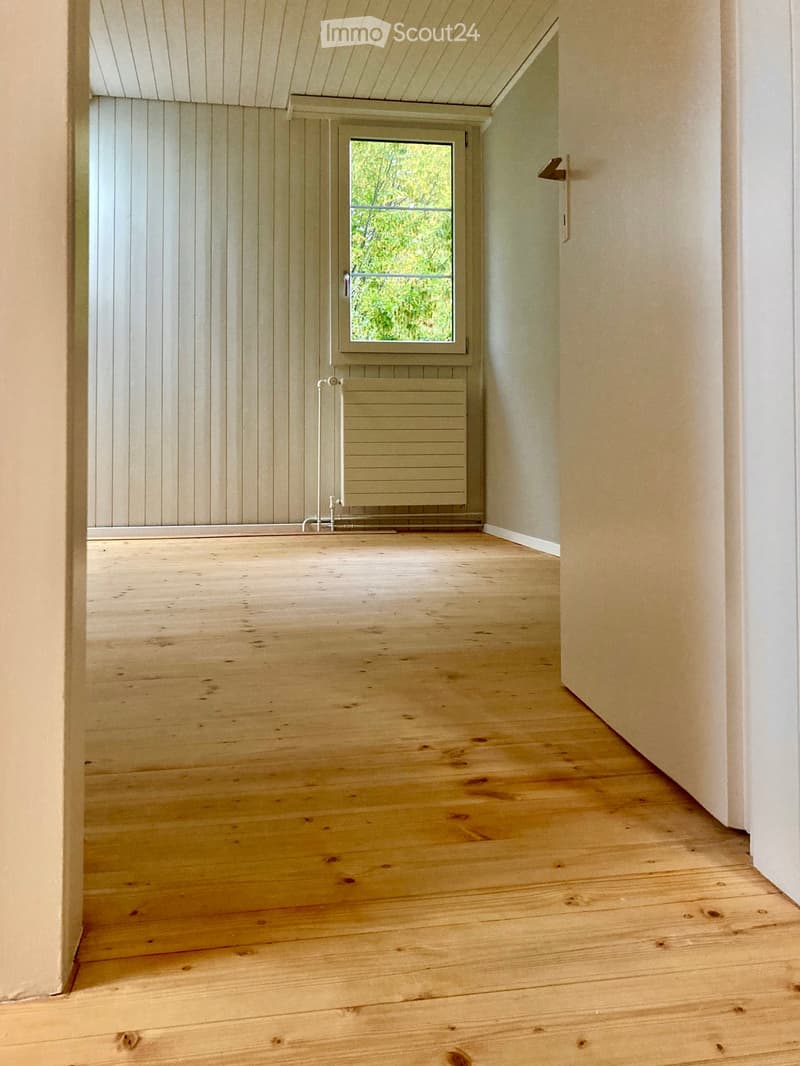 Empty hallway with wooden flooring, white walls, and a radiator. A window at the end allows natural light to enter.