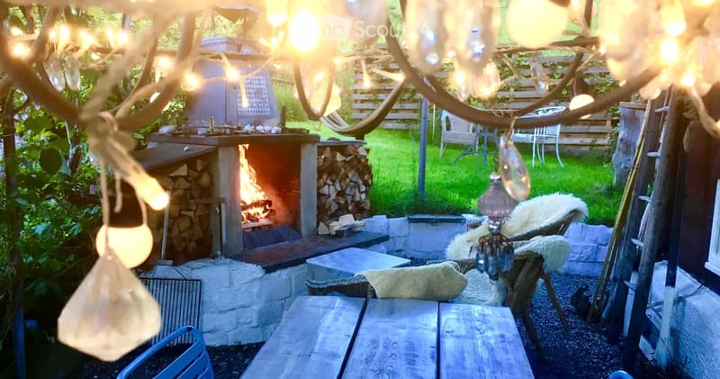 A cozy outdoor area with a stone fireplace, wooden tables, chairs, and string lights hanging from a tree branch. There is a stack of firewood and a grassy lawn in the background.