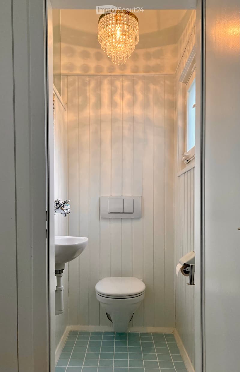 Compact bathroom with white paneled walls, teal tiled floor, a toilet, a sink with a modern faucet, and a decorative chandelier light fixture.