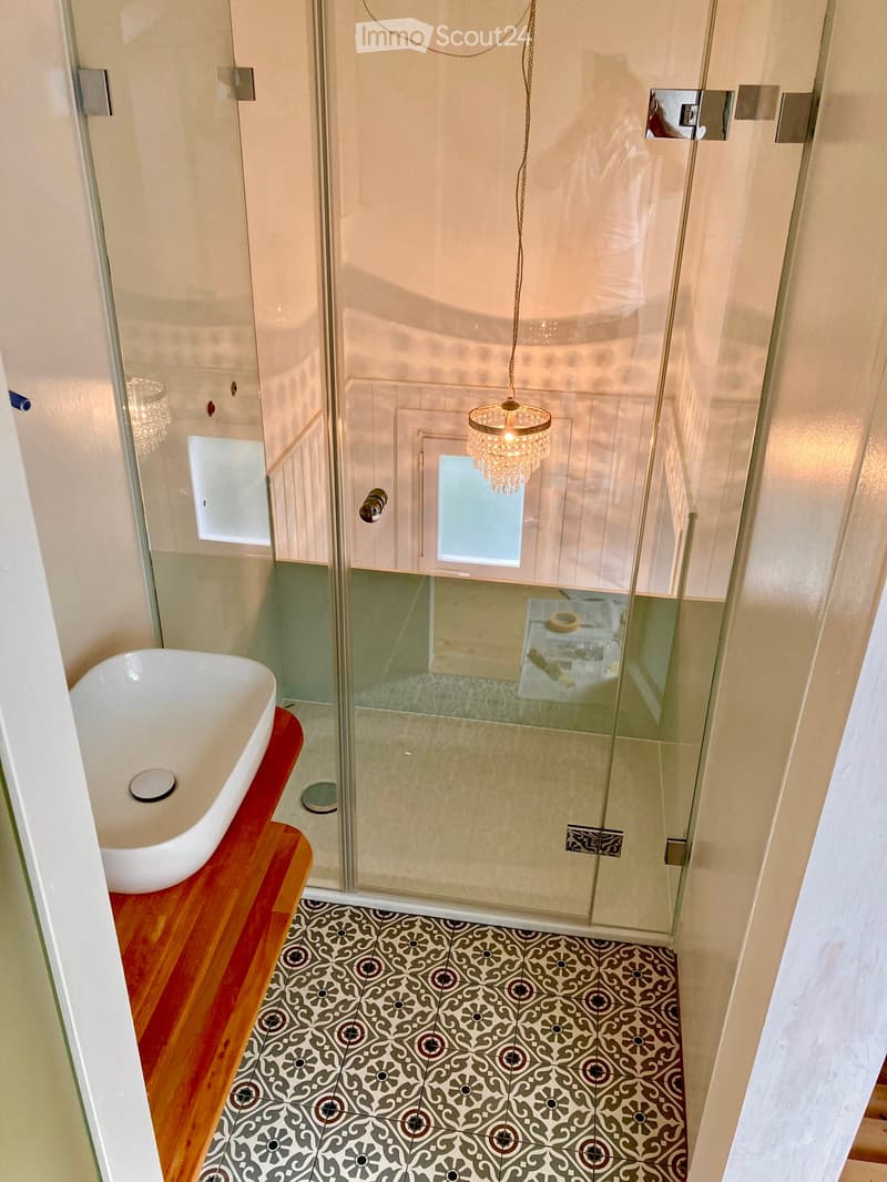 Elegant bathroom with a white bathtub on a wooden platform, a glass-enclosed shower with a chandelier, and patterned tiles on the floor.