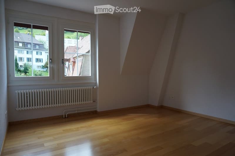 Empty room with wooden flooring, white walls, and large windows offering a view of neighboring houses and greenery. A radiator is visible below the windows.