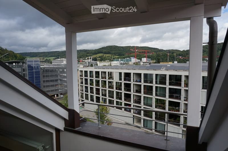 Balcony overlooking a modern urban area with buildings, greenery, and distant hills under a cloudy sky.