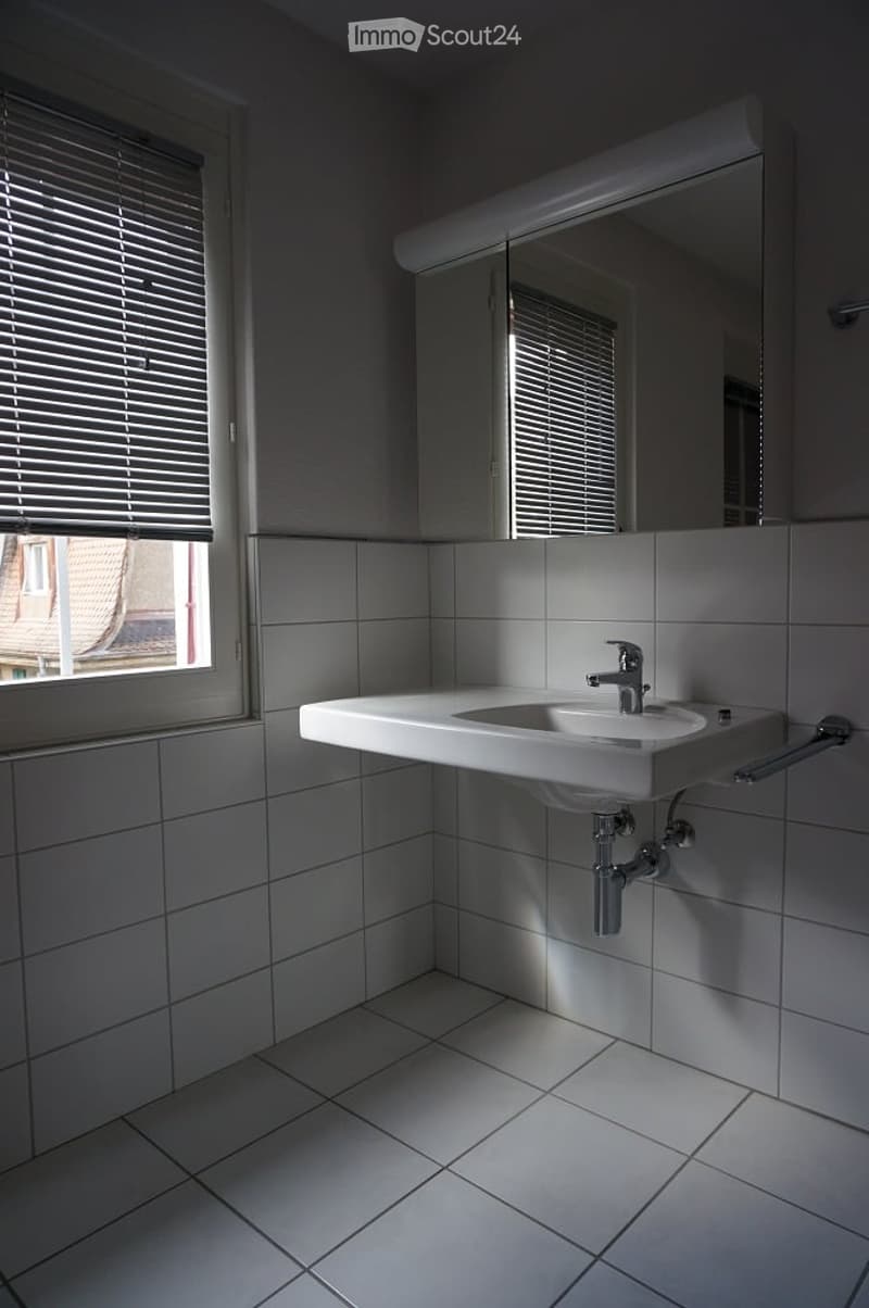 Modern bathroom with white tiled walls and floor, a single sink with a modern faucet, and a large mirror above it. The room has a window with blinds allowing natural light to enter.