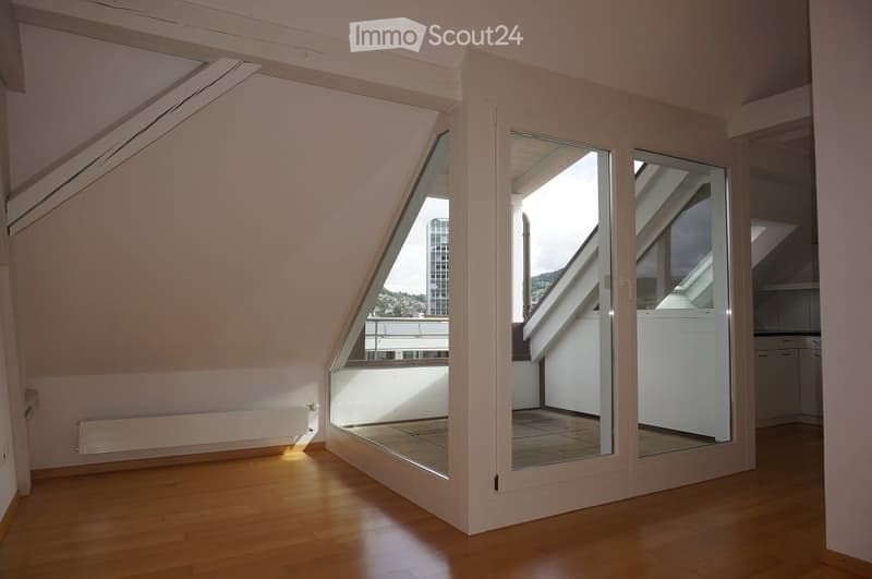 Bright, empty attic room with wooden flooring, large skylight windows, and a view of the city and surrounding landscape.