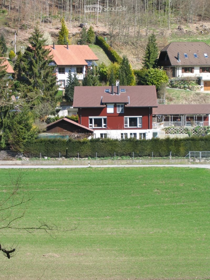 Residential houses with red-tiled roofs and white walls, surrounded by greenery and a grassy field.