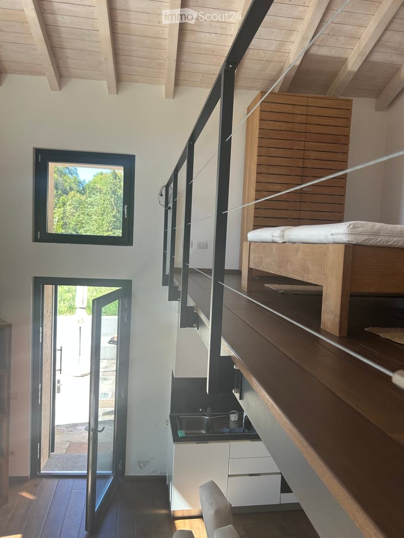 Modern interior with wooden staircase, open-plan living and sleeping area, large windows, and a small kitchenette.