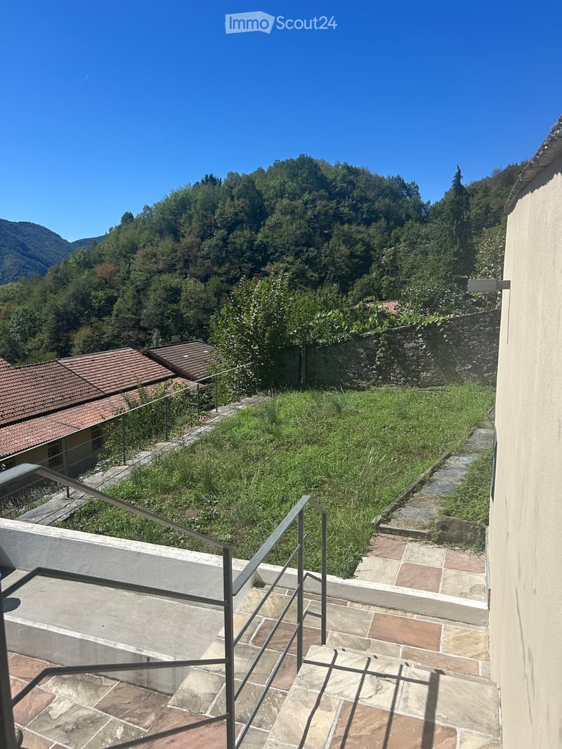 Balcony with stone steps, metal railing, and a view of a grassy area, stone wall, and forested hills in the background.