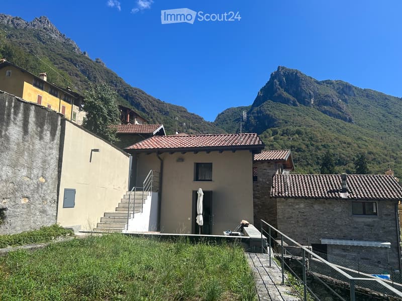 A two-story house with a tiled roof, stone and plaster exterior, and a small porch with an umbrella. The house is situated in a mountainous area with a staircase leading up to the entrance.
