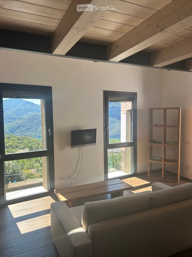 Modern living room with exposed wooden beams, large windows offering a scenic mountain view, a grey sofa, a wooden coffee table, a wooden shelf, and a wall-mounted television.