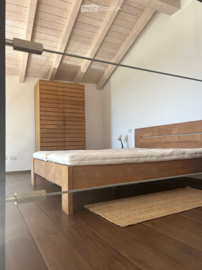 Modern bedroom with wooden bed frame, white mattress, nightstand, lamp, and small woven rug on wooden floor.