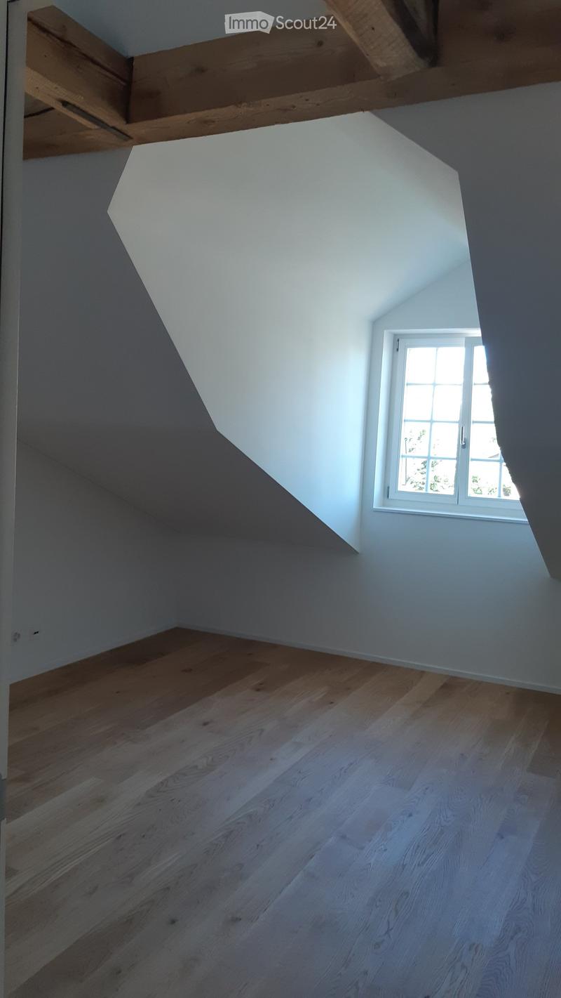 Empty attic space with wooden flooring, white walls, and a single window letting in natural light. Exposed wooden beams on the ceiling.