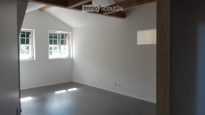 Empty room with white walls, grey floor, and three windows allowing natural light. Exposed wooden beams on the ceiling.