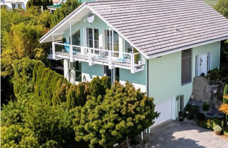 A two-story house with a light blue exterior, grey roof, and multiple large windows. Features include a balcony with outdoor seating, a small garden area, and a driveway.