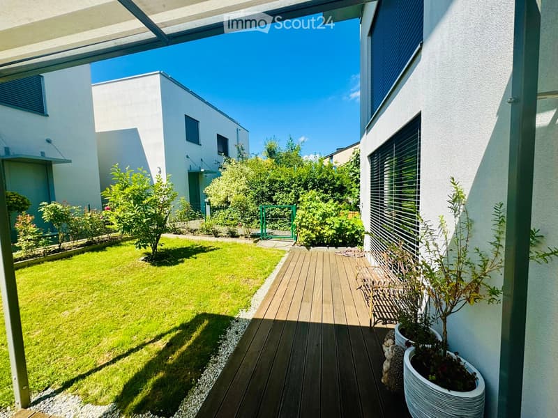Modern garden with a wooden deck, green lawn, potted plants, and a small bench. The area is enclosed by a white building with large windows and a green gate.