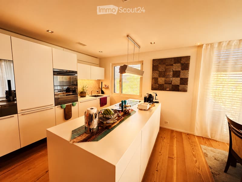Modern kitchen with white cabinets, a central island, and wooden flooring. Features include a sink, oven, coffee maker, and decorative items on the island.