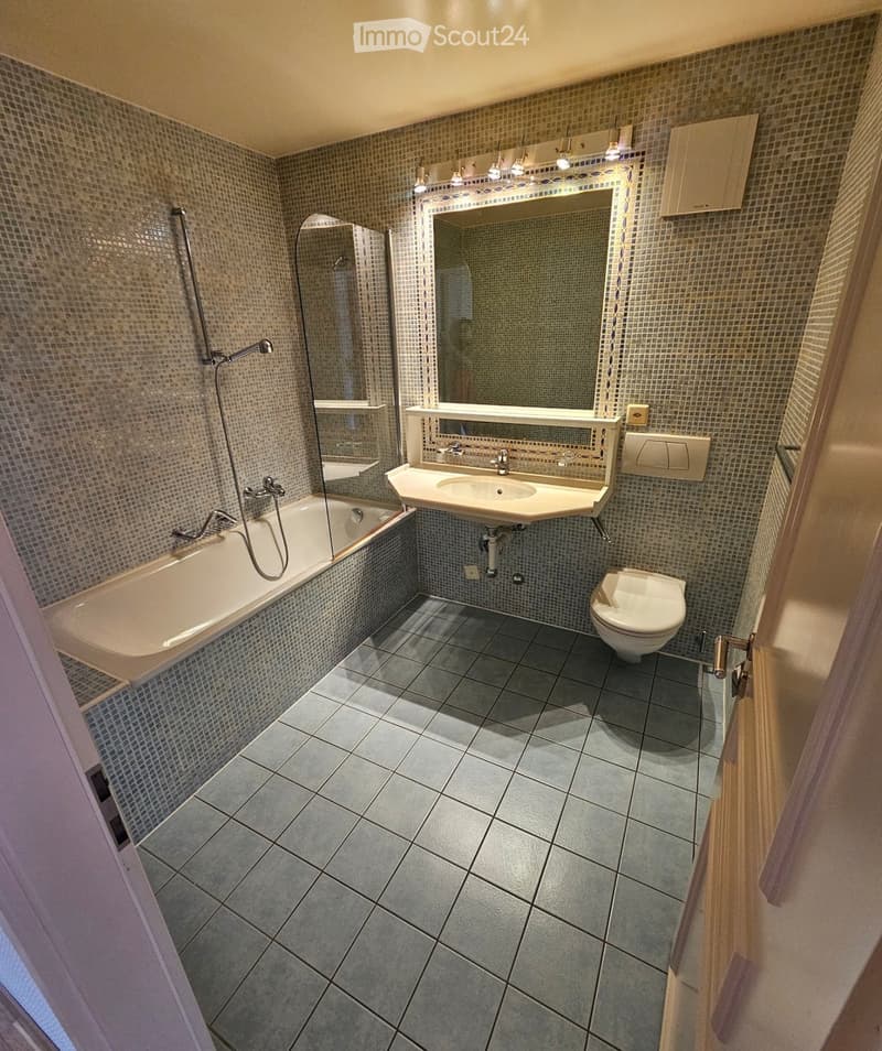 Modern bathroom with a bathtub, sink, toilet, and large mirror. The walls are tiled with small mosaic tiles, and the floor is covered with large grey tiles.