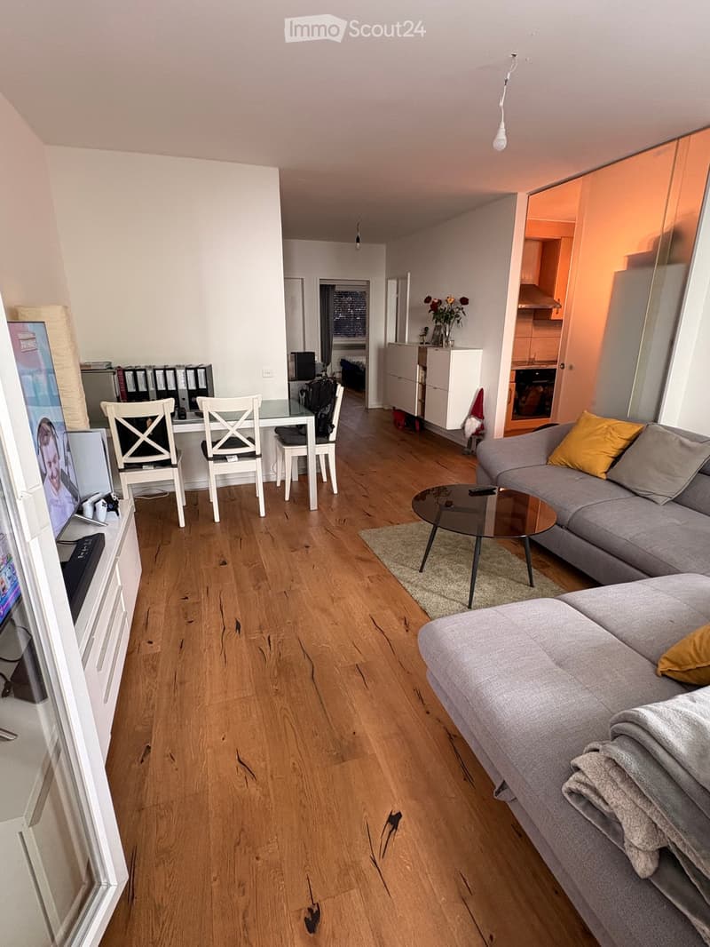 Living room with wooden flooring, grey sofa, coffee table, dining area with chairs and table, TV screen, and a small cabinet with a flower vase.