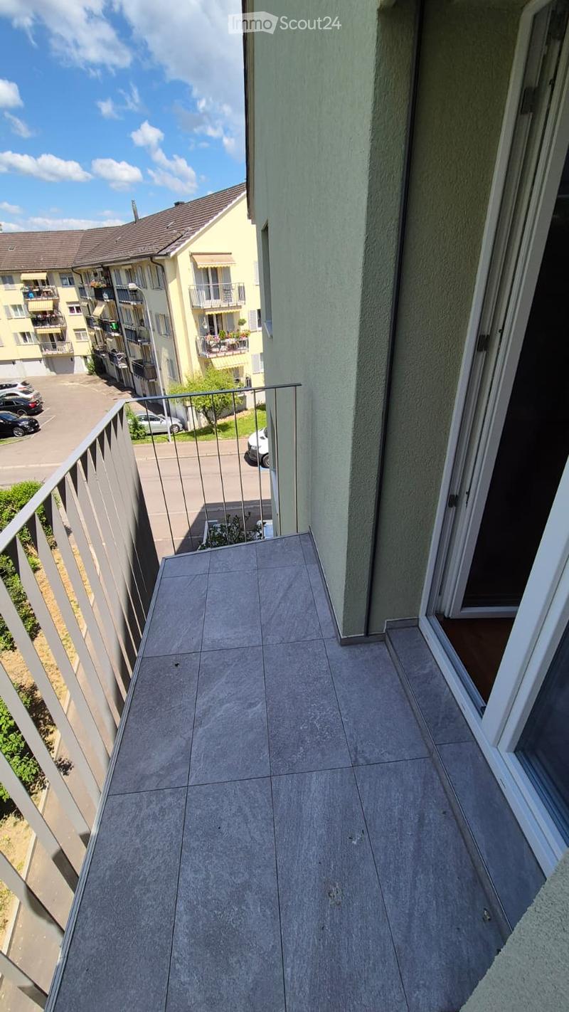 Balcony with grey tiled floor, metal railing, overlooking a residential area with parked cars and neighboring buildings.