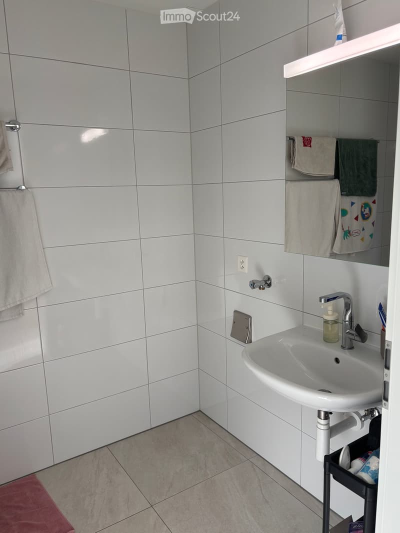 Modern bathroom with white tiled walls, a white sink with a chrome faucet, a mirror with a built-in shelf holding towels, and a small storage cart with toiletries.