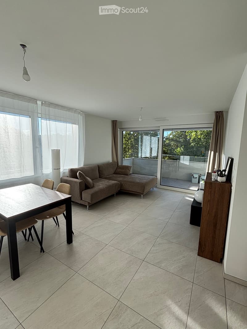 Modern living room with a grey sectional sofa, a dining table with chairs, large windows with curtains, and a balcony access through glass doors.