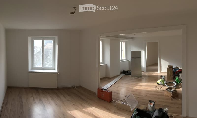 Empty room with wooden flooring, white walls, and large windows. Some construction materials and tools are visible, indicating ongoing renovation.